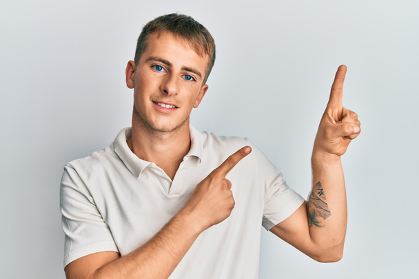 Jonge blanke man met casual witte polo glimlachend en kijkend naar de camera wijzend met twee handen en vingers naar de zijkant.  - Foto, afbeelding