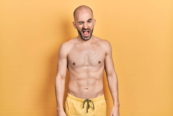 Junger glatzköpfiger Mann in Badebekleidung, der augenzwinkernd in die Kamera blickt, mit sexy Gesichtsausdruck, fröhlichem und glücklichem Gesicht.  - Foto, Bild