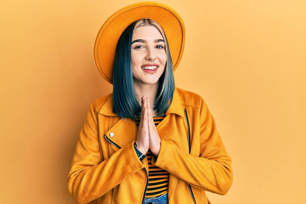 Fiatal, modern lány sárga kalapban és bőrdzsekiben, kéz a kézben imádkozva, bocsánatért esedezve, magabiztosan mosolyogva..  - Fotó, kép