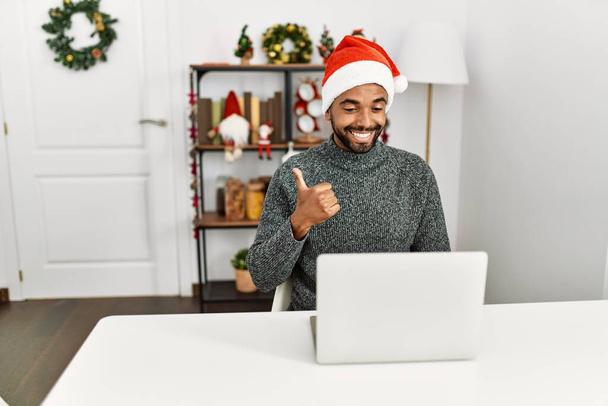 Joven hombre hispano con barba usando sombrero de navidad usando portátil sonriendo feliz y positivo, pulgar hacia arriba haciendo excelente y signo de aprobación  - Foto, Imagen