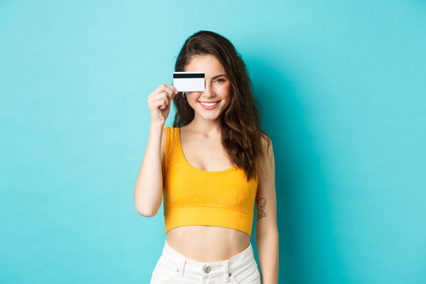 Młoda stylowa kobieta pokazuje plastikową kartę kredytową nad okiem, wygląda zadowolony i pewny siebie, uśmiecha się do kamery, stoi na niebieskim tle - Zdjęcie, obraz