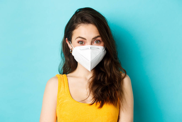 Covid-19, Coronavirus und soziale Distanzierung. Attraktive junge Frau mit Atemschutzmaske vor Coronavirus, Blick in die Kamera, blauer Hintergrund - Foto, Bild