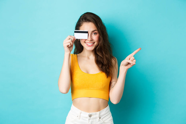 Stylish summer girl with credit card pointing aside, showing your logo on right side copy space, standing against blue background - Foto, imagen
