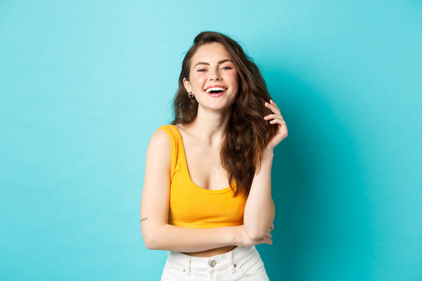 Summer and lifestyle concept. Portrait of attractive woman laughing and playing with her hair, standing in yellow cropped top, going on holiday, standing against blue background - Foto, Imagem