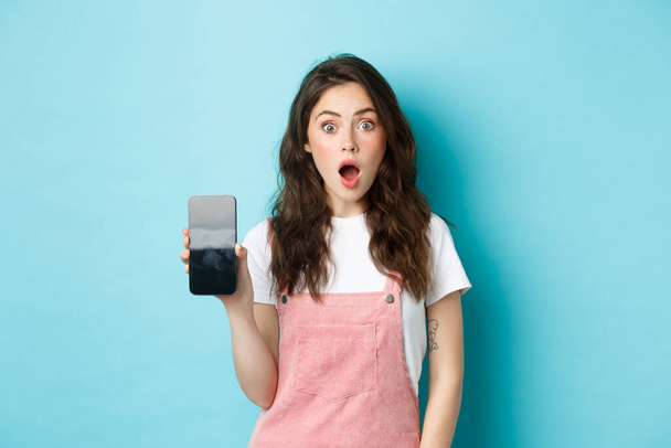 Shocked beautiful girl drop jaw and show empty smartphone screen, demonstrate your app or online store on blank phone display, standing against blue background - Фото, изображение