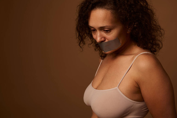 Εξαντλημένη φοβισμένη γυναίκα με σφραγισμένο στόμα φαίνεται απελπισμένα κάτω, στέκεται τρία τέταρτα σε ένα σκούρο μπεζ φόντο με χώρο για κείμενο. Κοινωνική έννοια του τερματισμού της βίας κατά των γυναικών - Φωτογραφία, εικόνα