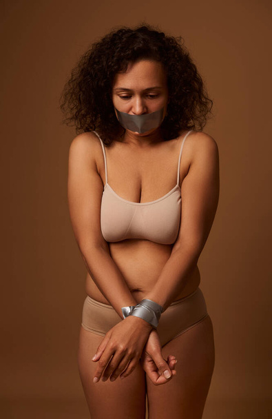 Portrait of frightened desperate mixed race Hispanic woman with sealed mouth looking down with hopelessness, isolated on dark beige background. International Day to Eliminate Violence against Women. - Photo, image