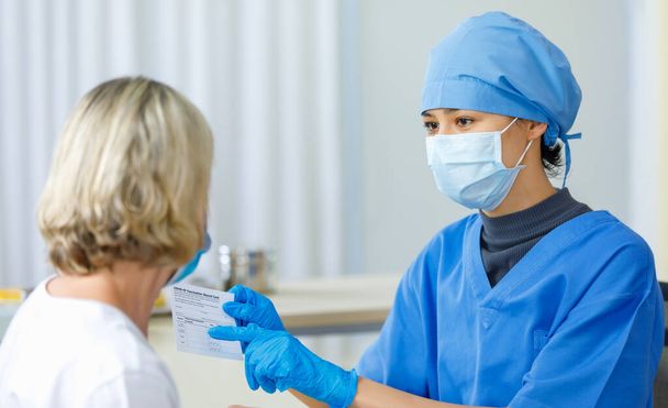 Młoda lekarka w niebieskim szpitalnym mundurze maska na twarz i gumowe rękawiczki pokazują jak wyjaśnić Covid-19 karta szczepień poświadcza blond pacjentce rasy kaukaskiej. - Zdjęcie, obraz