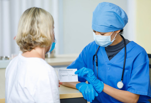 Doctor in blue hospital uniform face mask and rubber gloves with stethoscope send and explain Covid-19 vaccination record card certificate to Caucasian female patient in blurred foreground. - Fotoğraf, Görsel