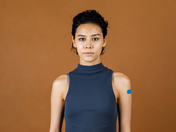 Retrato Closeup estúdio caneca tiro de bonito bonito sexy paciente feminino stand sorrindo mostrando bandagem de gesso azul em seu ombro depois de receber vacinação Coronavirus Covid-19 na parede marrom da frente. - Foto, Imagem