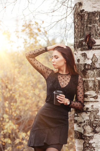 Portrait of pretty young woman of Slavic appearance in dark dress with cup of coffee in autumn, standing next to birch tree against background of an autumn Park. Golden fall. Copy space - Foto, afbeelding