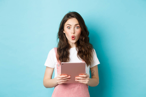 Impressed cute girl say wow, stare excited at camera, holding digital tablet over chest, standing in spring clothes against blue background - 写真・画像
