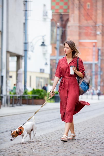 νεαρή γυναίκα που βγάζει βόλτα ένα σκύλο στην πόλη πίνοντας καφέ - Φωτογραφία, εικόνα