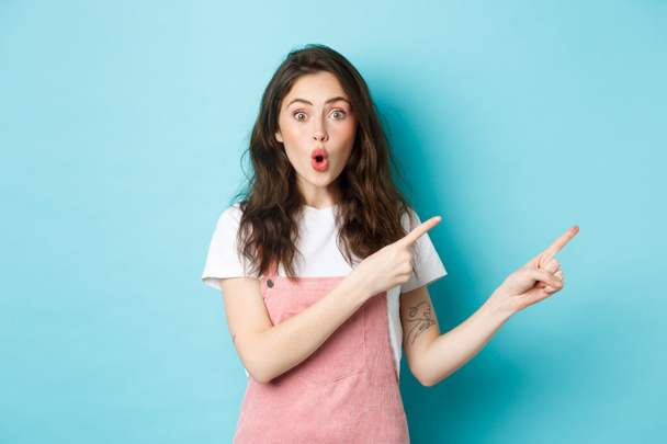 Impressed brunette girl pointing fingers right at logo banner, staring with excitement and surprised face, telling big news, showing promo offer, blue background - Zdjęcie, obraz
