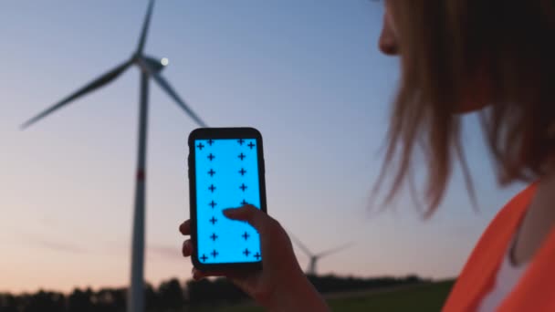 クロマキー付きのスマートフォンを持ち、日没時に風力タービンの背景にスクロールダウンジェスチャーを作る女性エンジニア. - 映像、動画