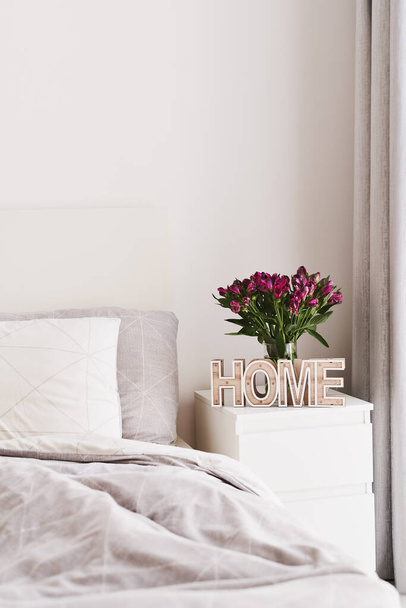 Κρεβατοκάμαρα διακόσμηση: λευκό κρεβάτι με γκρι κρεβάτι και δύο μαξιλάρια, λευκό κομοδίνο με ξύλινη ταμπέλα "σπίτι" και φρέσκα λουλούδια Αλστρομέρια σε βάζο. Ελάχιστος σκανδιναβικός εσωτερικός σχεδιασμός - Φωτογραφία, εικόνα
