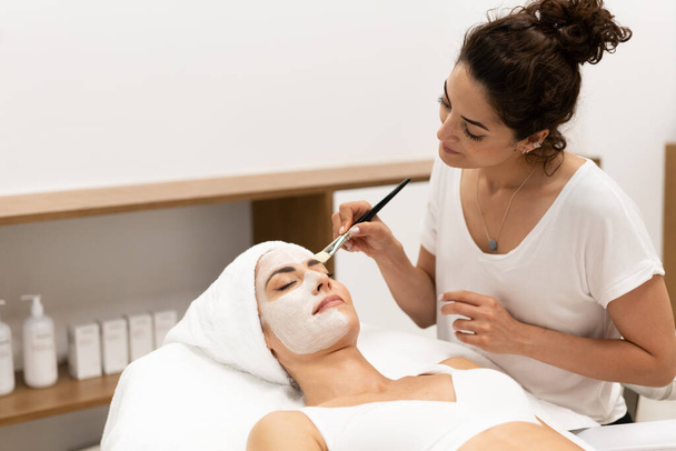 Estética aplicando uma máscara no rosto de uma mulher de meia-idade no centro de bem-estar moderno. - Foto, Imagem