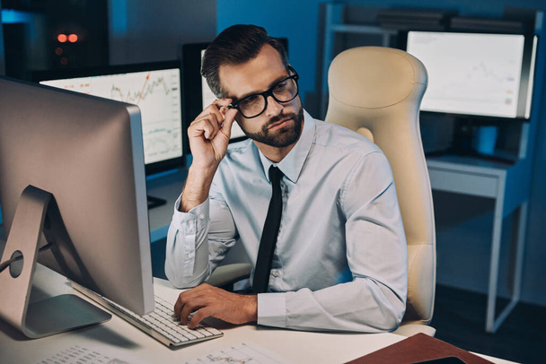 Confident young man adjusting his eyeglasses while staying late in the office - Фото, зображення
