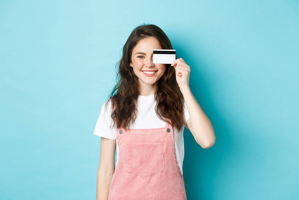 Cute young woman showing plastic credit card on eye and smiling, shopping with it, standing over blue background - Photo, Image