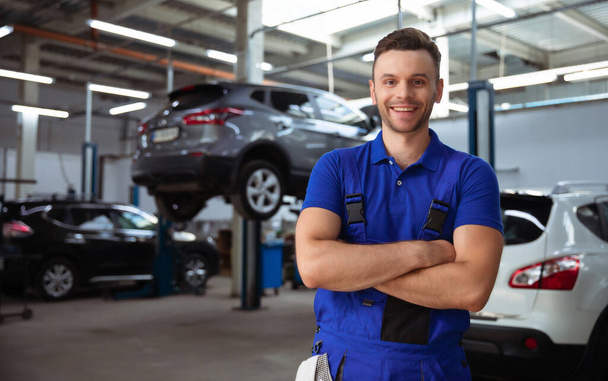 Confident handsome young and experienced car repair worker in work overalls posing against the background of lifted cars in a car service - 写真・画像