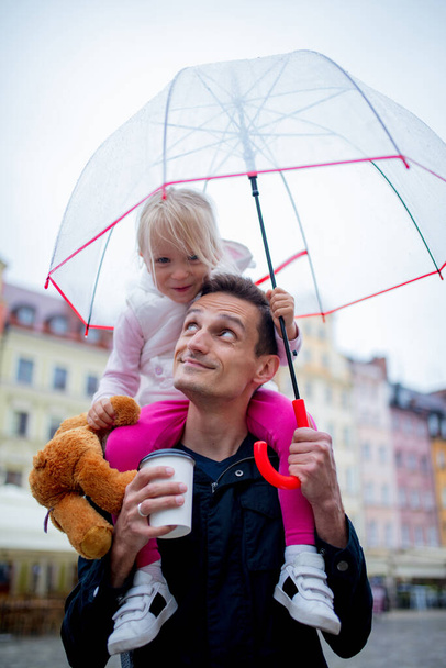 little girl with umbrella sits on her father's shoulders in the city - Photo, Image