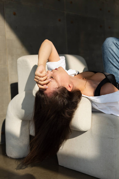 Jolie jeune femme avec un gros sein posant sur une chaise blanche en studio, mur en acier derrière, portant un jean et une chemise blanche, lumière naturelle du coucher de soleil chaud - Photo, image