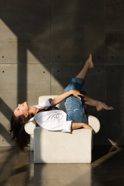 Hübsche junge Frau mit großer Brust posiert auf weißem Stuhl im Studio, Stahlwand dahinter, trägt Jeans und weißes Hemd, natürliches warmes Sonnenuntergangslicht - Foto, Bild