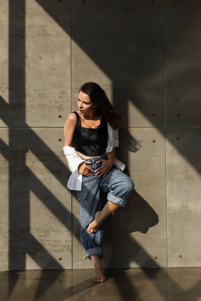 Красивая молодая женщина с большой грудью позирует в студии, за стальной стеной на заднем плане, в джинсах и белой рубашке, естественный теплый закат - Фото, изображение