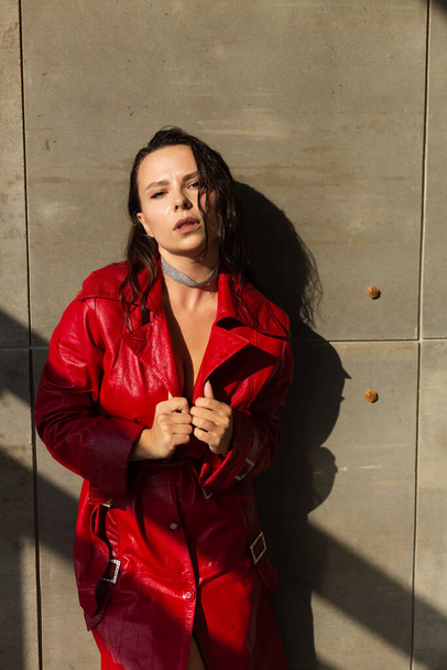 Csinos fiatal nő nedves haj pózol a stúdióban, visel bőr piros köpeny és fényes nyaklánc, természetes meleg naplemente fény, álló fal - Fotó, kép