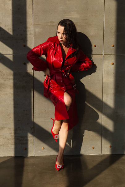 Mujer joven y bonita con el pelo mojado posando en el estudio, con capa de cuero rojo y collar brillante, luz natural cálida puesta del sol, de pie por la pared - Foto, Imagen