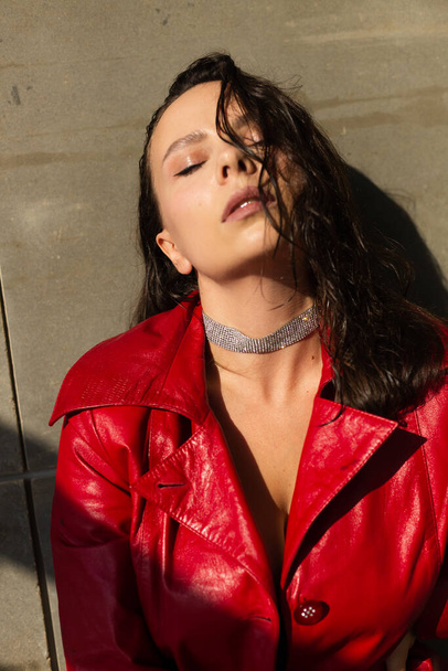 Pretty young woman with wet hair posing in studio, wearing leather red cloak and shiny necklace, natural warm sunset light, standing by wall - Foto, Imagen