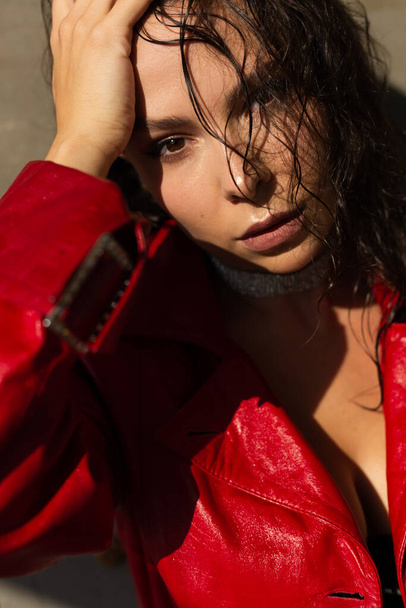 Ładna młoda kobieta z mokrymi włosami pozująca w studio, ubrana w skórzaną czerwoną pelerynę i błyszczący naszyjnik, naturalne ciepłe światło zachodu słońca, stojąca przy ścianie - Zdjęcie, obraz