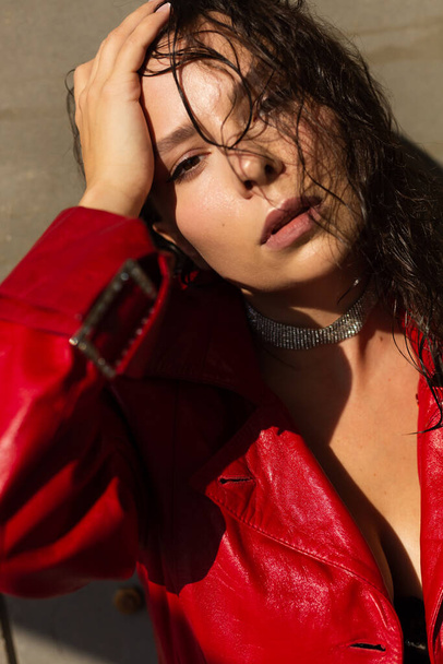 Красивая молодая женщина с мокрыми волосами позирует в студии, одета в кожаный красный плащ и блестящее ожерелье, естественный теплый закат света, стоя у стены - Фото, изображение