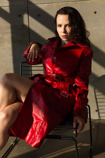 Mujer joven y bonita con el pelo mojado posando en el estudio, con capa de cuero rojo y collar brillante, luz natural cálida puesta del sol, se sienta en la silla - Foto, Imagen