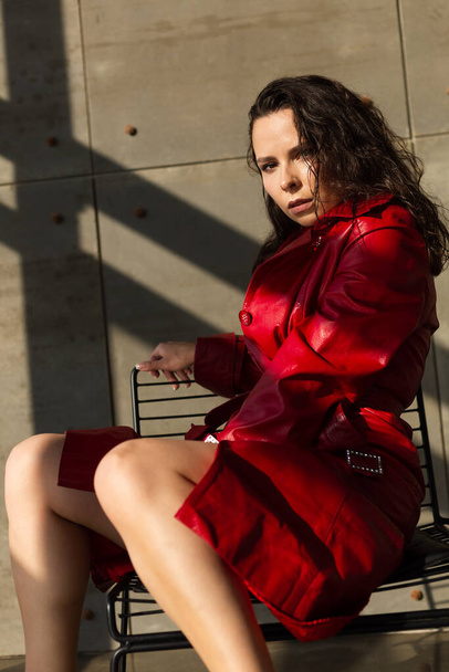 Pretty young woman with wet hair posing in studio, wearing leather red cloak and shiny necklace, natural warm sunset light, sits on chair - Valokuva, kuva