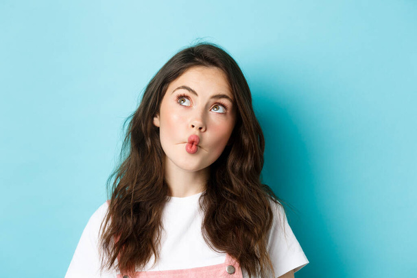 Close up portrait of funny young woman with clean skin and curly hairstyle, pucker lips thoughtful, looking at upper left corner dreamy, imaging something, standing over blue background - Photo, Image