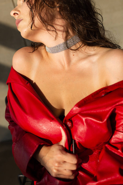 Красивая молодая женщина с мокрыми волосами позирует в студии, одета в кожаный красный плащ и блестящее ожерелье, естественный теплый закат света, сидит на стуле - Фото, изображение