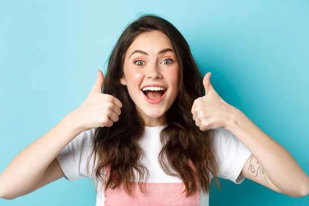 close-up van opgewonden brunette vrouw tonen duimen omhoog en zeggen ja, eens, geven positieve feedback, aanbevelen goed product, lof reclame, blauwe achtergrond - Foto, afbeelding