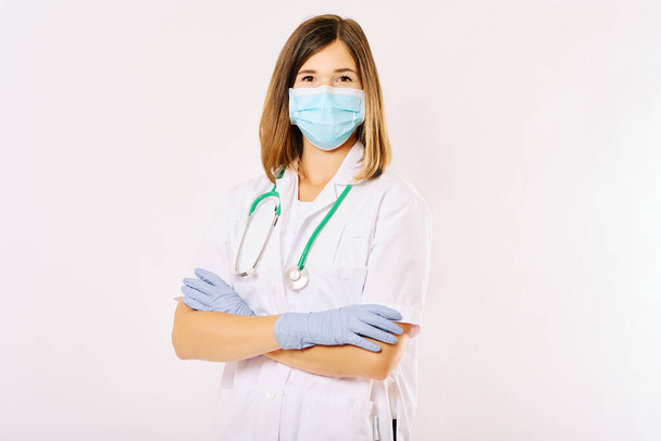 Studio portrait of young female doctor posing on white background, wearing facial mask and gloves, arms crossed - Foto, afbeelding