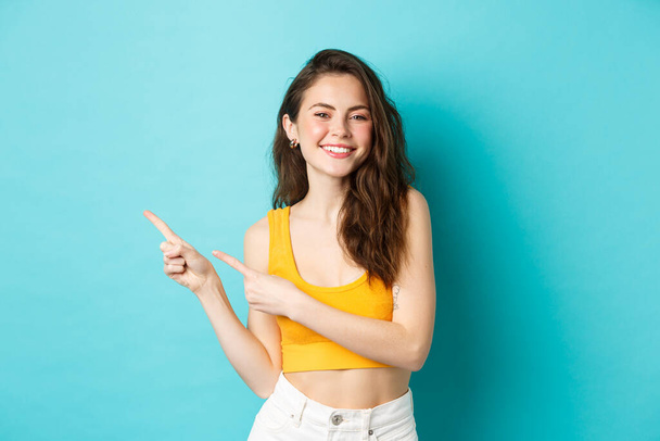 Belle jeune femme regardant heureux, souriant avec des dents, invitant à vérifier promo événement, pointant les doigts gauche à l'espace de copie, debout sur fond bleu - Photo, image