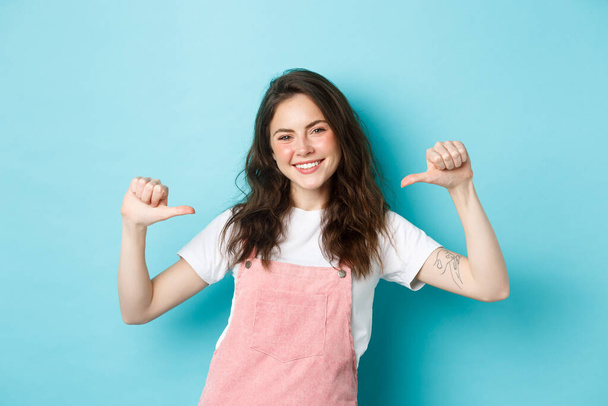 Young smiling woman pointing at herself, looking confident and happy, self-promoting, talking about her, personal achievement, standing over blue background - Photo, Image
