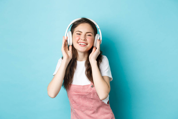 Cute caucasian girl in spring outfit, listening music in headphones, smiling pleased at camera, standing over blue background - Photo, Image
