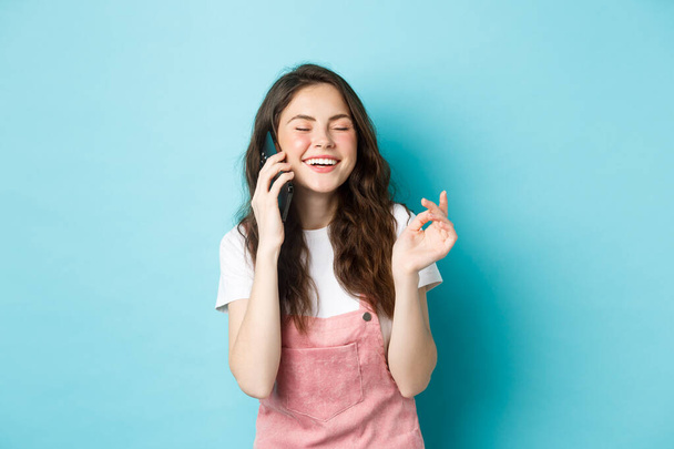 Retrato de chica linda y elegante riendo mientras chatea en el teléfono, sosteniendo el teléfono inteligente cerca de la oreja y sonriendo feliz, tener una conversación informal, de pie sobre el fondo azul - Foto, Imagen