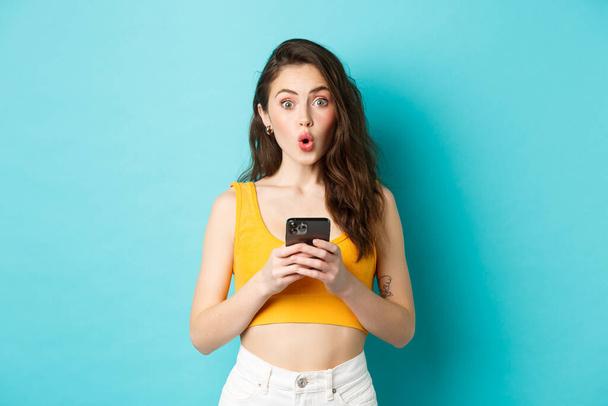Looks interesting. Intrigued pretty girl holding smartphone, staring excited and saying wow after reading online advertisement, standing over blue background - Foto, immagini