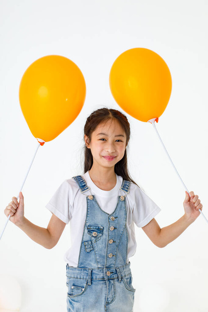 Portrait close up isolated studio shot of young pretty cute long black hair preschooler girl in jeans denim skirt overalls standing smiling look at camera holding two orange balloon sticks in hands. - Foto, Imagen