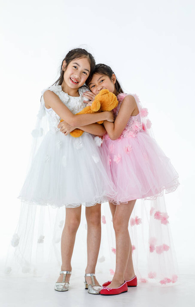 Portrait close up isolated studio shot of two little Asian ballerina kids in pink and white roses flowers ballet dress smiling laughing happily while playing fighting over orange teddy bear doll. - Foto, afbeelding