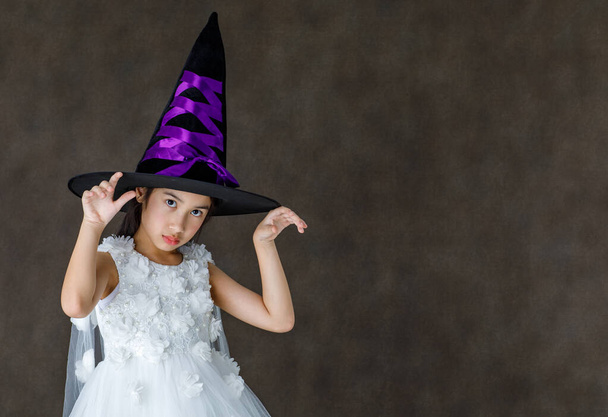 Portrait close up studio shot of Asian cute little girl in witch dress costume with tall hat stand smile look at camera ready to play trick or treat on Halloween festival. - Фото, изображение