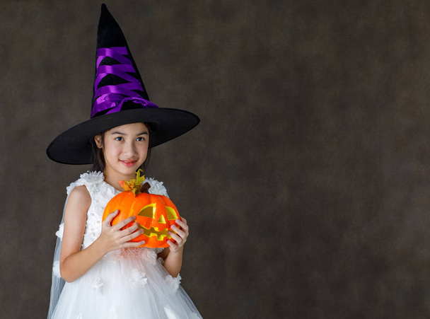 Portrait close up studio shot of Asian cute little girl in witch dress costume with tall hat stand smile look at camera hold orange pumpkin in hand ready to play trick or treat on Halloween festival. - 写真・画像