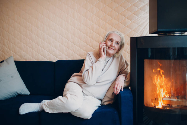 Elderly gray haired woman sitting on sofa and talking smartphone in living room with fireplace - Photo, Image