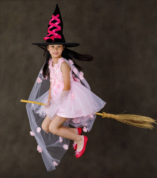 Портрет студии снимок маленького милого азиатского ребенка в розовом костюме ведьмы в высокой черной шляпе посмотрите на камеру, позируя летающий жест верхом на волшебной метле колдовства на Хэллоуин традиционный фестиваль. - Фото, изображение